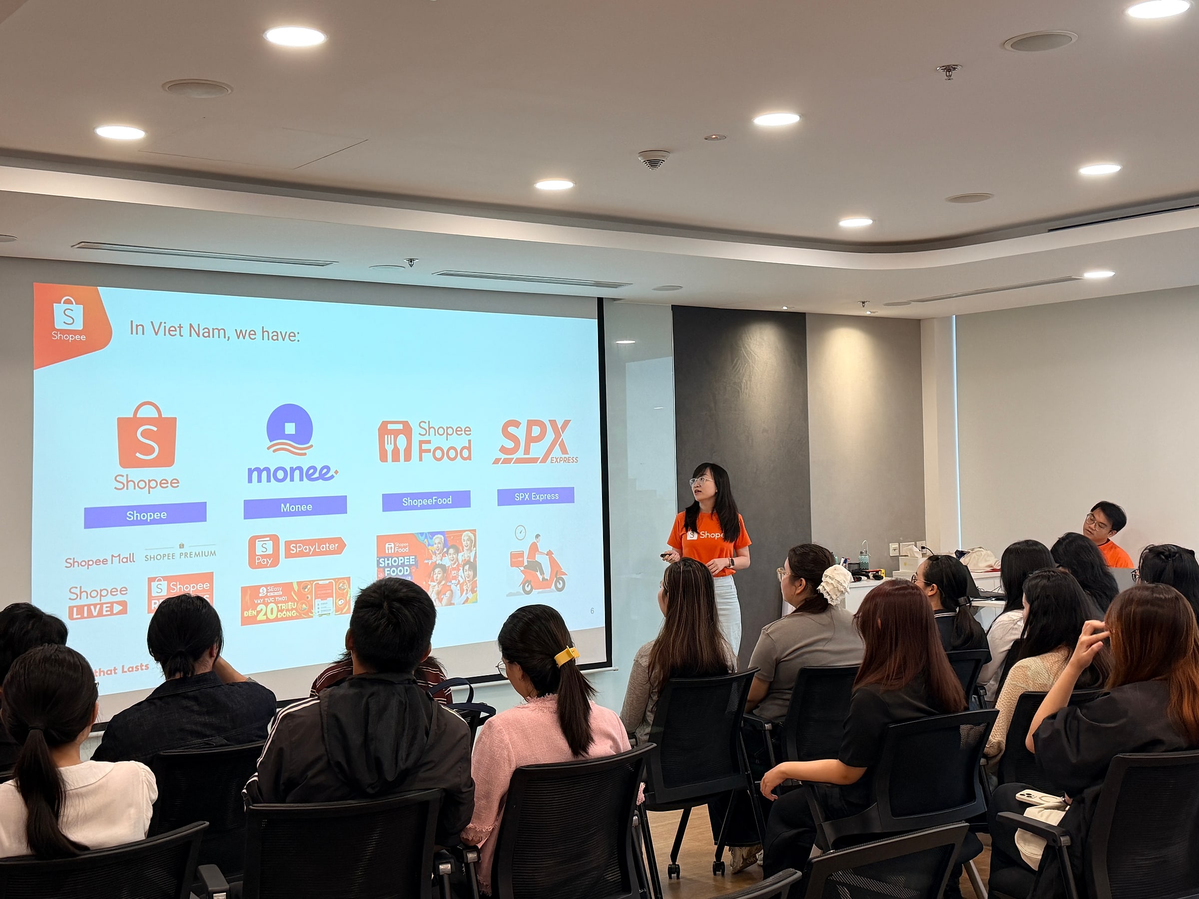 Sinh viên IU ngồi nghe giới thiệu trong phòng họp của Shopee, màn chiếu hiển thị Shopee, Monee, ShopeeFood và SPX Express