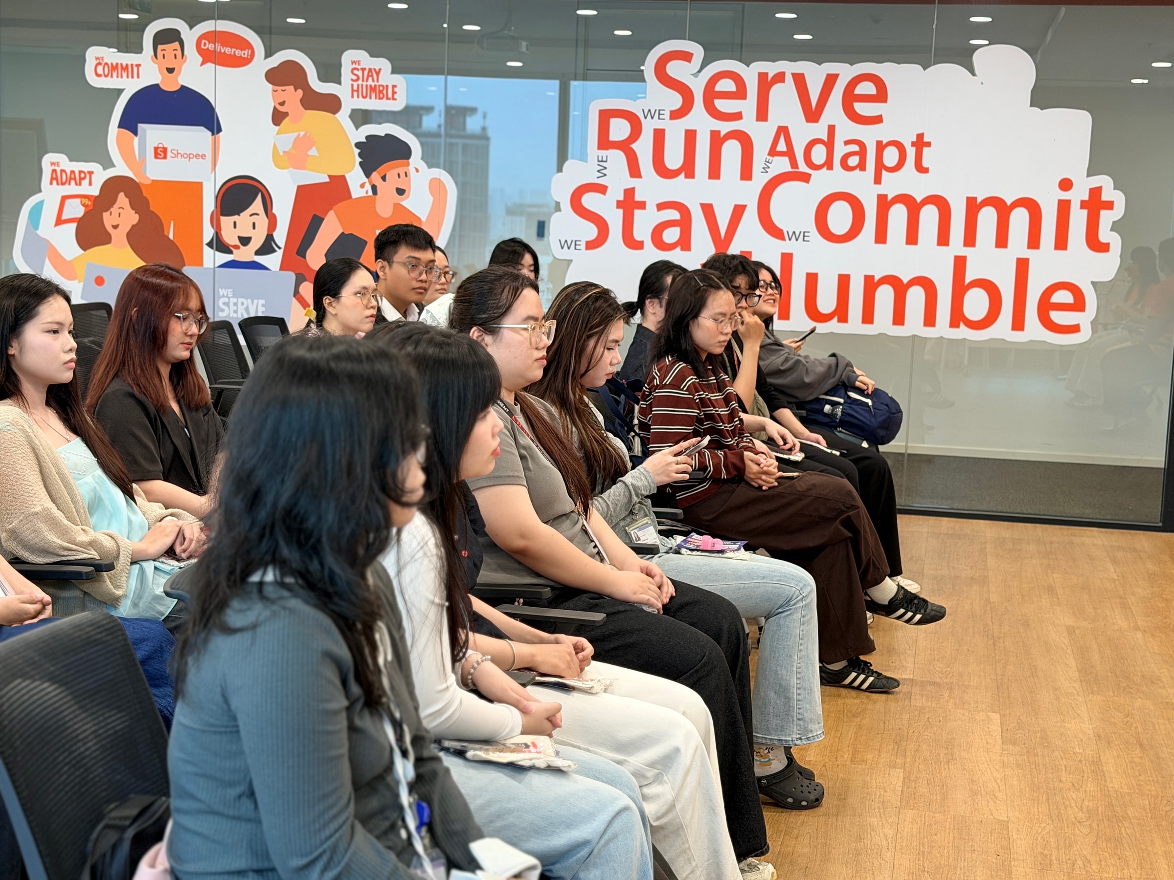 Sinh viên IU ngồi trước bức tường in các giá trị Serve, Run, Adapt, Stay Humble, Commit của Shopee