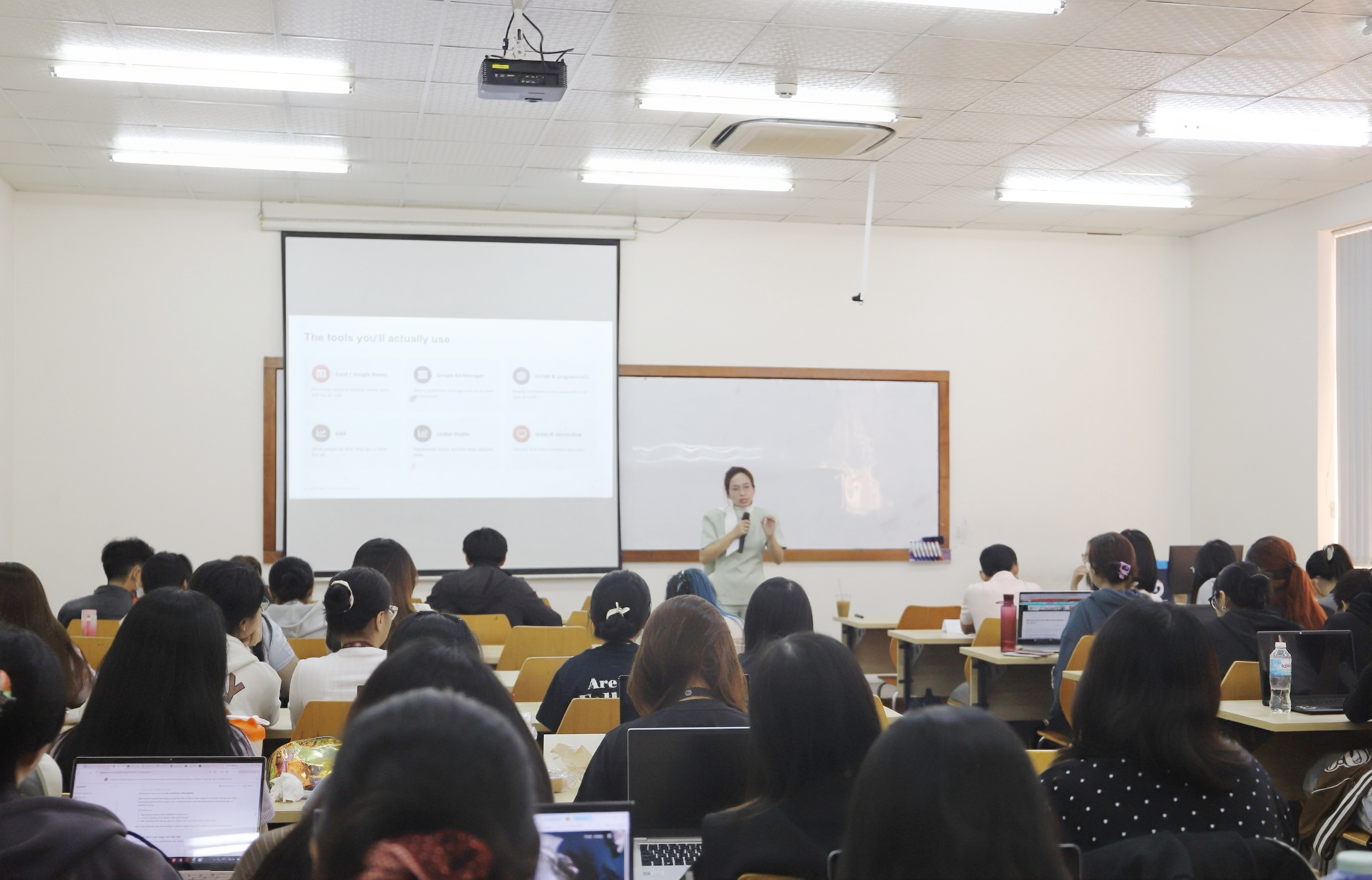 An Nguyễn cầm micro đứng trước lớp BA275IU, màn chiếu hiển thị slide về các công cụ trong ngành quảng cáo