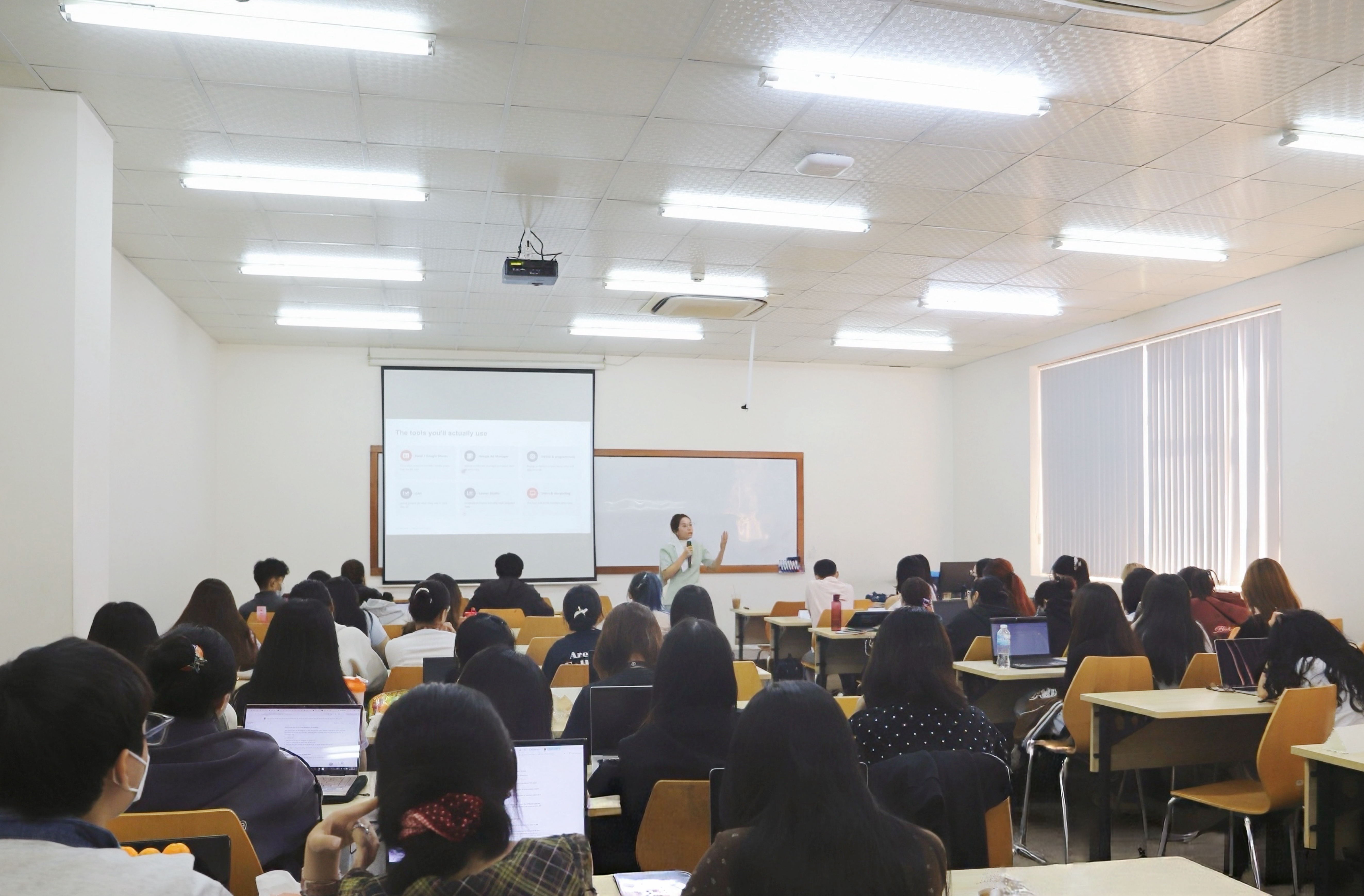 Toàn cảnh phòng học nhìn từ cuối lớp, sinh viên mở laptop theo dõi phần trình bày